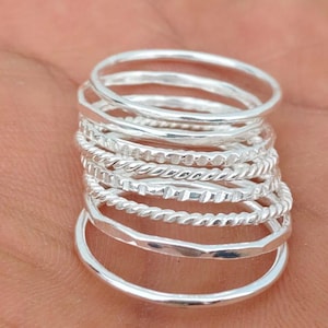 Può includere: Una pila di anelli d'argento in vari design, tra cui fasce martellate, intrecciate e lisce. Gli anelli sono esposti sulla mano di una persona, mostrando le diverse trame e stili dei gioielli.