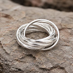 May include: A silver ring composed of multiple interlocking bands, creating a layered effect. The ring is displayed on a textured, gray-brown stone surface, highlighting its metallic shine. The design is simple yet elegant, suitable for everyday wear.