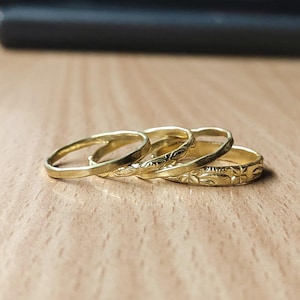 May include: Four gold rings, each with a unique design. One ring is smooth, while the others feature etched patterns of leaves and flowers. The rings are stacked on a light brown wooden surface.