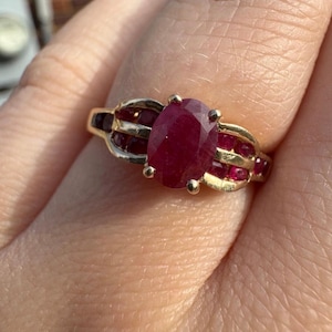 May include: A gold ring featuring a large, oval-cut ruby as the centerpiece, flanked by smaller, rectangular rubies. The ring has a detailed, ornate design with a warm, golden hue. The ruby is a deep, rich red color.