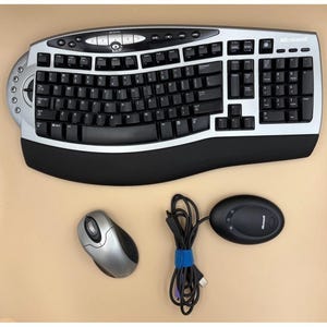 Puede incluir: Un conjunto de teclado y ratón Microsoft plateado y negro. El teclado ergonómico tiene un diseño curvo con teclas negras y un borde plateado. También se incluye un ratón plateado y un ratón ovalado negro.