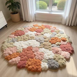 Peut inclure: Un tapis rond texturé avec un motif en forme de nuages. Le tapis présente une variété de couleurs, notamment le blanc, le rose, l'orange et le gris. Le tapis est fait d'un matériau doux et est posé sur un sol en bois.