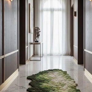 Puede incluir: Un pasillo con paneles de madera oscura, molduras doradas y suelo de mármol. Una alfombra grande y texturizada en tonos verdes y blancos se asemeja a un paisaje de musgo. El espacio está iluminado por apliques de pared y luz natural de una gran ventana.