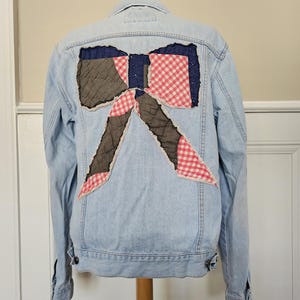 May include: Light blue denim jacket with a large bow design on the back. The bow is made of quilted gray, navy blue, and red and white gingham fabric. The jacket has a classic collar, button closures, and pockets.