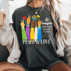 Puede incluir: Sudadera gris oscuro con un gráfico colorido de manos que muestran signos de paz y sostienen un corazón, un símbolo de paz y una flor. El texto "PEACE & LOVE" e "Imagine all the people living life in peace" también están impresos en la prenda.