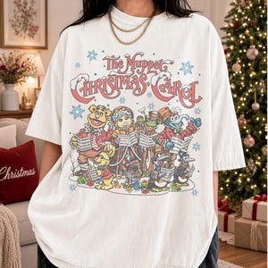 May include: A white short-sleeve t-shirt featuring a colorful graphic print of The Muppet Christmas Carol characters singing from songbooks, surrounded by blue snowflakes and the movie title in red script lettering across the top.