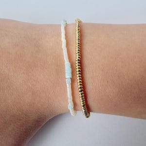 May include: Two bracelets are displayed. One is a delicate strand of small, gold beads. The other is a white beaded bracelet with light blue square beads interspersed throughout. The bracelets are worn on a wrist.