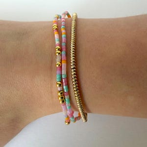 May include: Three bracelets on a wrist. Two are beaded with pastel colors and gold accents. The third is a gold-colored, textured bangle. The bracelets are stacked together. The beads are pink, blue, orange, and yellow.