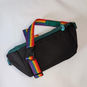 Pode incluir: Cinto de cintura preto com parte superior verde-azulado e uma alça arco-íris. A palavra "LEITERS" está impressa em verde na alça. A pochete tem uma fivela preta e um zíper turquesa.