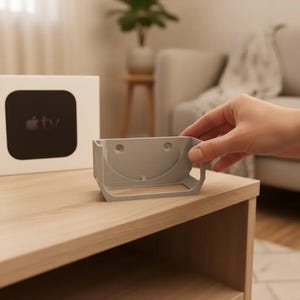 以下が含まれることがあります： 手で持たれた、曲線的なデザインのライトグレーのApple TVマウント。 マウントは明るい木製の表面にあり、Apple TVボックスの隣にあります。 Apple TVボックスは白で、黒い画面とAppleのロゴが付いています。