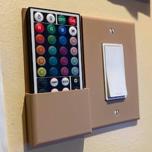 May include: A light brown remote control holder mounted next to a light switch. The remote has multiple colored buttons and is designed to control lights. The light switch is white and rectangular.