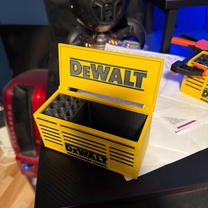 Könnte beinhalten: Eine gelb-schwarze Werkzeugkiste mit dem "DEWALT"-Logo auf dem Deckel. Die Box ist geöffnet und zeigt einen unterteilten Innenraum. Die Worte "GUARANTEED TOUGH" sind auf der Seite der Box aufgedruckt.