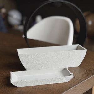 May include: Two white textured rectangular planters, one larger than the other, sit on a wooden surface. A black circular object is in the background. The planters have a textured surface and are empty.