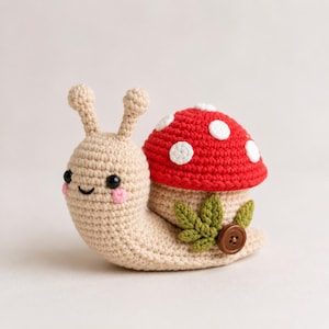 Könnte beinhalten: Eine beige gehäkelte Schneckenfigur mit einem roten Pilzhut mit weißen Punkten. Die Schnecke hat schwarze Perlenaugen, rosa gestickte Wangen und einen grünen gehäkelten Blattakzent an der Seite, abgerundet mit einem kleinen braunen Knopf.