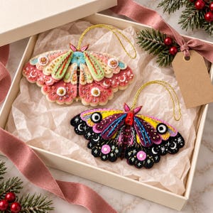 Könnte beinhalten: Zwei Motten-Ornamente aus Filz mit aufwendigen Stickereien und goldenen Aufhängeschlaufen. Eine Motte zeigt Rosa-, Creme- und Grüntöne mit Mondsichel-Details. Die zweite Motte ist schwarz, violett und gelb mit Stern- und Mondstickmustern.