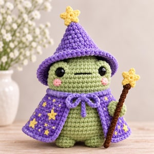 Könnte beinhalten: Eine hellgrüne, gehäkelte Froschfigur mit einem violetten Zaubererhut und einem passenden violetten Umhang, der mit kleinen gelben Sternen verziert ist. Der Frosch hat schwarze Augen, rosa Wangen und hält einen braunen Zauberstab mit gelbem Stern.