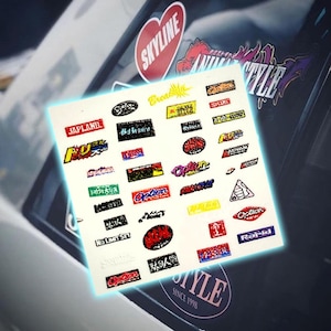Puede incluir: Una hoja de pegatinas con varios logotipos y textos, incluyendo "SKYLINE", "JAPLAND" y "Option". Las pegatinas son de varios colores y formas, con un enfoque en la marca automotriz. El fondo es una ventana de coche.