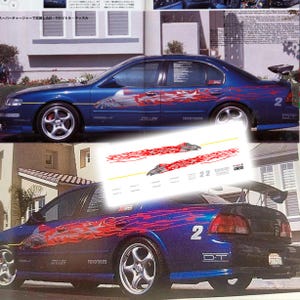 May include: A blue sedan with red flame decals and the number 2 on the rear. The car has aftermarket wheels and a rear spoiler. The text "STILLEN" and "TOYO TIRES" are visible on the car's body. A smaller image shows decals with the text "22 toucan".