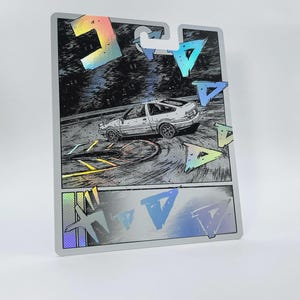 Könnte beinhalten: Eine rechteckige Karte mit einer schwarz-weißen Manga-Illustration eines weißen Schrägheckautos, das auf einer Strecke driftet. Das Design enthält holografische geometrische Formen und japanische Katakana-Schriftzeichen sowie eine Stanzung.