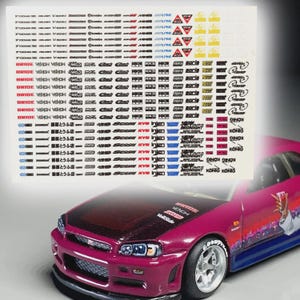 May include: A sheet of racing decals featuring logos and text such as "BRIDE," "HKS," and "MIDNIGHT." A deep pink model car is in the foreground, with a black bonnet and a graphic on the side.