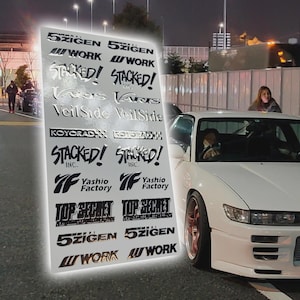 May include: A sheet of car decals featuring various brand names and logos, including "5ZIGEN," "WORK," and "STACKED!" The decals are in black and silver, with a white car in the background.