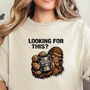 Könnte beinhalten: Beiges T-Shirt mit dem Text "LOOKING FOR THIS?" und einem Cartoon-Sasquatch, der ein 10-mm-Objekt hält. Die Grafik ist in Braun- und Beigetönen gehalten. Das Model trägt eine blaue Jeans.