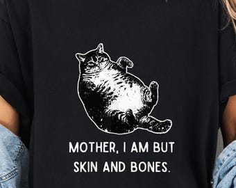 May include: Black t-shirt featuring a white illustration of a cat lying on its back. The text "MOTHER, I AM BUT SKIN AND BONES" is printed below the cat. The shirt is worn with a denim jacket and shorts.