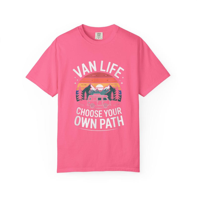 May include: A bright pink t-shirt with the words "VAN LIFE" above a graphic of a camper van, mountains, and trees. Below the graphic, the text reads "CHOOSE YOUR OWN PATH". The design features a sunset color scheme.