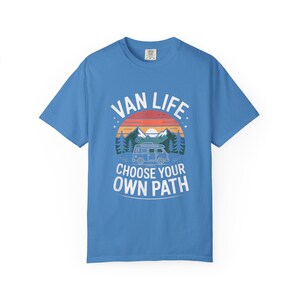 May include: A light blue t-shirt with the words "VAN LIFE" above a graphic of a van, mountains, and trees. Below the graphic, the text reads "CHOOSE YOUR OWN PATH". The design features a sunset color scheme.