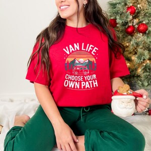 May include: A red t-shirt with the words "VAN LIFE CHOOSE YOUR OWN PATH" printed on the front. The graphic includes a camper van, mountains, and a sunset. The person is wearing green pants and holding a Santa mug.
