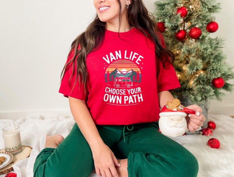 May include: A red t-shirt with the words "VAN LIFE CHOOSE YOUR OWN PATH" printed on the front. The graphic includes a camper van, mountains, and a sunset. The person is wearing green pants and holding a Santa mug.
