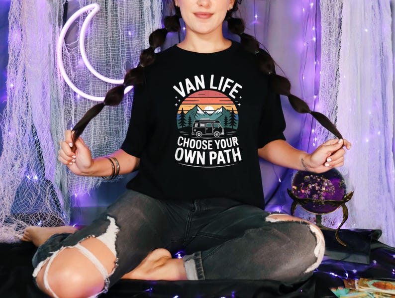 May include: Black t-shirt with the text "VAN LIFE CHOOSE YOUR OWN PATH" in white. The graphic features a van, mountains, and a sunset. The shirt is worn by a person.
