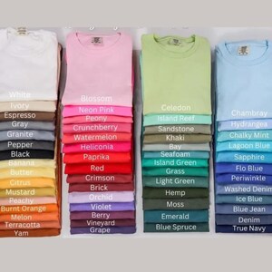 May include: A display of folded t-shirts in various colors, including white, pink, green, and blue. Each stack is labeled with the color name, such as "Blossom," "Celedon," and "Chambray." The shirts are neatly arranged, showcasing a range of hues.