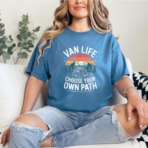 May include: A blue t-shirt with the words "VAN LIFE" above a graphic of a camper van, mountains, and a sunset. The text "CHOOSE YOUR OWN PATH" is below the graphic. The shirt is worn by a person sitting on a white couch.