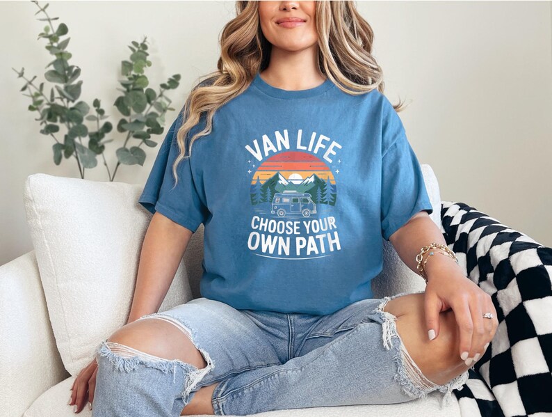 May include: A blue t-shirt with the words "VAN LIFE" above a graphic of a camper van, mountains, and a sunset. The text "CHOOSE YOUR OWN PATH" is below the graphic. The shirt is worn by a person sitting on a white couch.