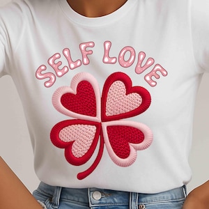 May include: White t-shirt with a four-leaf clover design, each leaf a heart shape in red and pink embroidery. The words "SELF LOVE" are above the clover in pink bubble letters. A casual, comfortable top.
