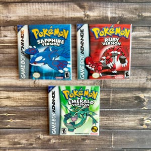 May include: Three Game Boy Advance video game cartridges: "Pokémon Sapphire Version" with a blue creature, "Pokémon Ruby Version" with a red creature, and "Pokémon Emerald Version" with a green creature. The games are on a wooden surface.