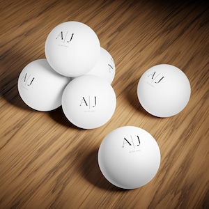 May include: A collection of white spherical balls, each featuring the black printed letters A | J and the date 20.08.2027. The smooth, matte-finish spheres are arranged in a cluster, showcasing a consistent minimalist design across every individual ball.