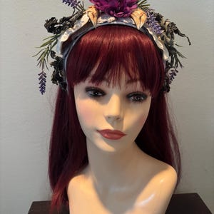 May include: A decorative headband featuring a central arrangement of a deep purple flower, surrounded by dried foliage and lavender sprigs. The headband's base is a light blue fabric, creating a whimsical and naturalistic aesthetic.