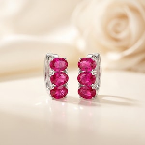 May include: A pair of silver hoop earrings, each featuring three oval-cut, vivid red gemstones. The earrings are set against a soft, blurred background, emphasising the jewellery's elegant design and colour.
