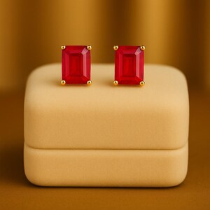 May include: Gold stud earrings featuring rectangular, bright red gemstones. The earrings are presented on a beige jewellery box against a warm, golden-brown backdrop. The studs have small, round gold accents.
