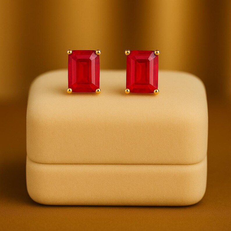 May include: Gold stud earrings featuring rectangular, bright red gemstones. The earrings are presented on a beige jewellery box against a warm, golden-brown backdrop. The studs have small, round gold accents.
