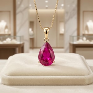 May include: A gold necklace featuring a pear-shaped, faceted deep pink gemstone pendant. The pendant is suspended from a delicate gold box chain, showcasing a polished gold bail that connects the vibrant, teardrop-cut stone to the chain.