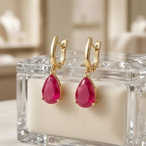 May include: A pair of gold-toned drop earrings featuring pear-shaped, vibrant pink-red gemstones suspended from hinged clasps. Each clasp is clearly stamped with the S925 hallmark, indicating sterling silver construction with a gold-tone finish.