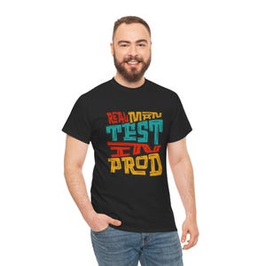 Könnte beinhalten: Ein schwarzes kurzärmeliges T-Shirt mit einem Grafikdruck, der den Text "REAL MAN TEST IN PROD" in einer stilisierten, abgenutzten Schriftart zeigt. Die Buchstaben sind in den Farben Rot, Gelb und Türkis in vier horizontalen Zeilen angeordnet.