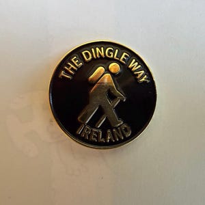 May include: A round, black pin with a gold border and lettering. The pin features the words "THE DINGLE WAY IRELAND" in gold. A gold figure of a person hiking with a walking stick is in the center.
