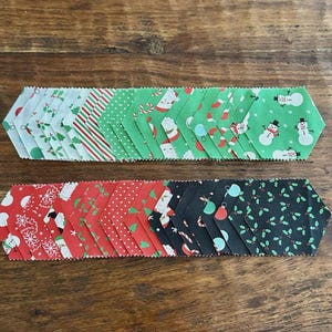 May include: Two rows of Christmas-themed fabric strips arranged on a wooden surface. The top row features green, white, and red patterns with Santa, candy canes, and snowmen. The bottom row has red, black, and white fabrics with similar festive designs.