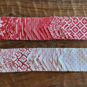May include: Two rows of red and white fabric hexagon shapes with various patterns, including floral, striped, and geometric designs. The fabric pieces are arranged on a wooden surface, showcasing a variety of quilting fabric.