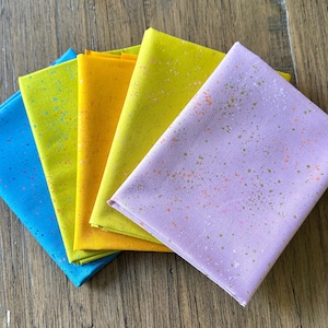 May include: A stack of colorful fabric squares in shades of blue, yellow, and purple. Each fabric square is speckled with contrasting colors. The fabrics are folded and arranged on a wooden surface.