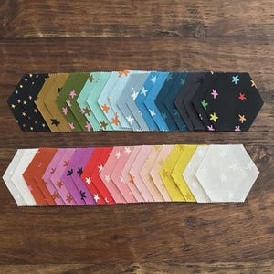 May include: Arrangement of colorful fabric hexagon pieces. The top row features dark colors with star patterns, while the bottom row showcases lighter shades with star designs. The fabrics are arranged on a wooden surface.